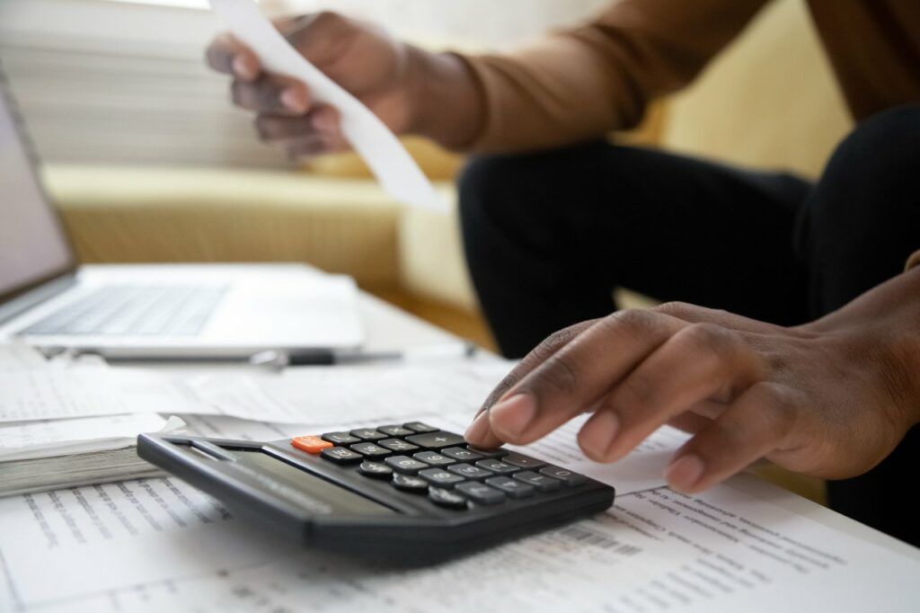 A man types numbers into a calculator while referring to several documents and a laptop.