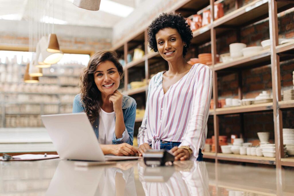 Two women smile at the camera with a laptop and a card reader on the table and shelves in the background.