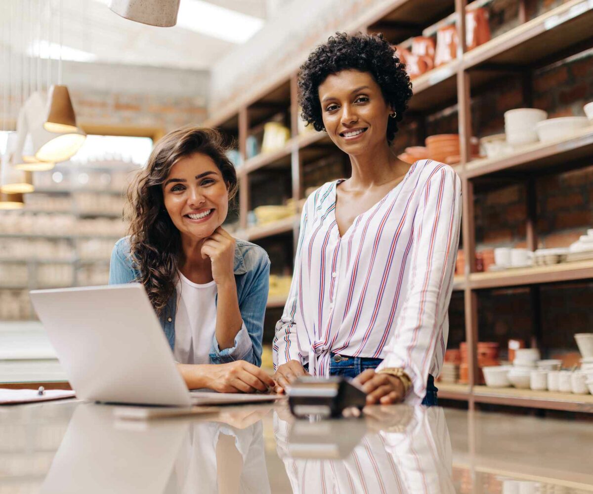 Two women smile at the camera with a laptop and a card reader on the table and shelves in the background.