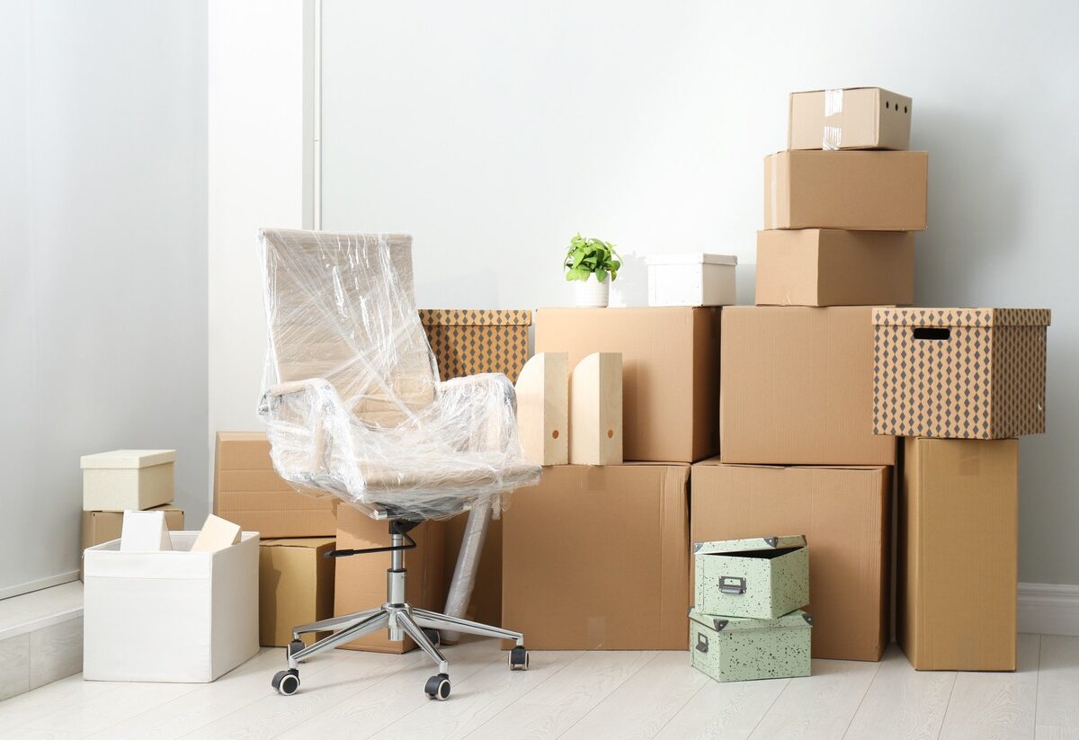 A stack of moving boxes and a plastic-wrapped desk chair sit against a wall