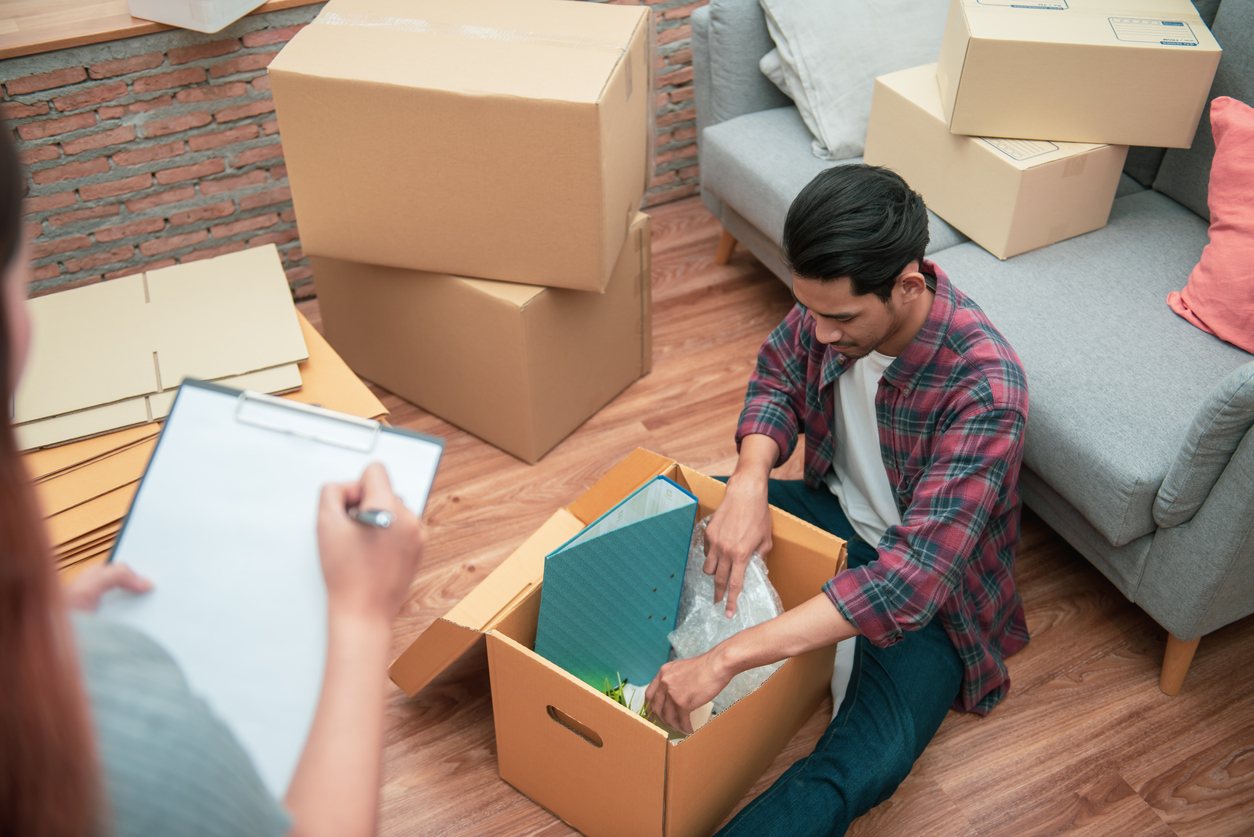 A man sits on the floor and packs a box with belongings, while his partner writes on a checklist.