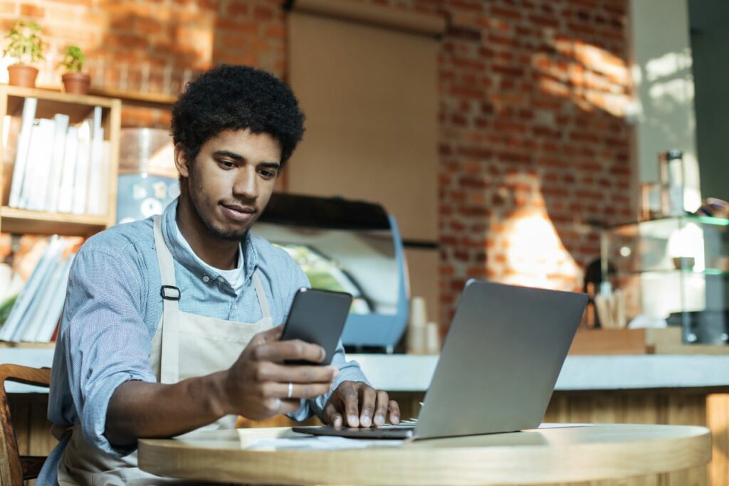 A young small business owner sits at a table, conducting business on his phone and laptop.