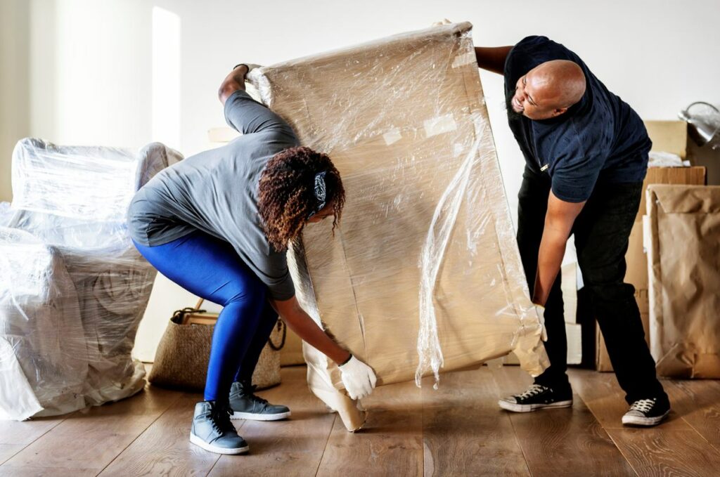 Two people wear gloves as they carefully lift a dresser wrapped in plastic.