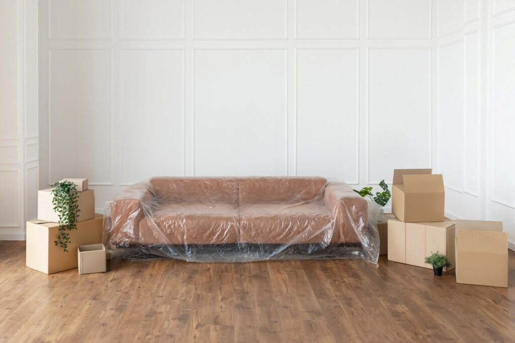 A sofa wrapped in plastic surrounded by boxes in a white room.