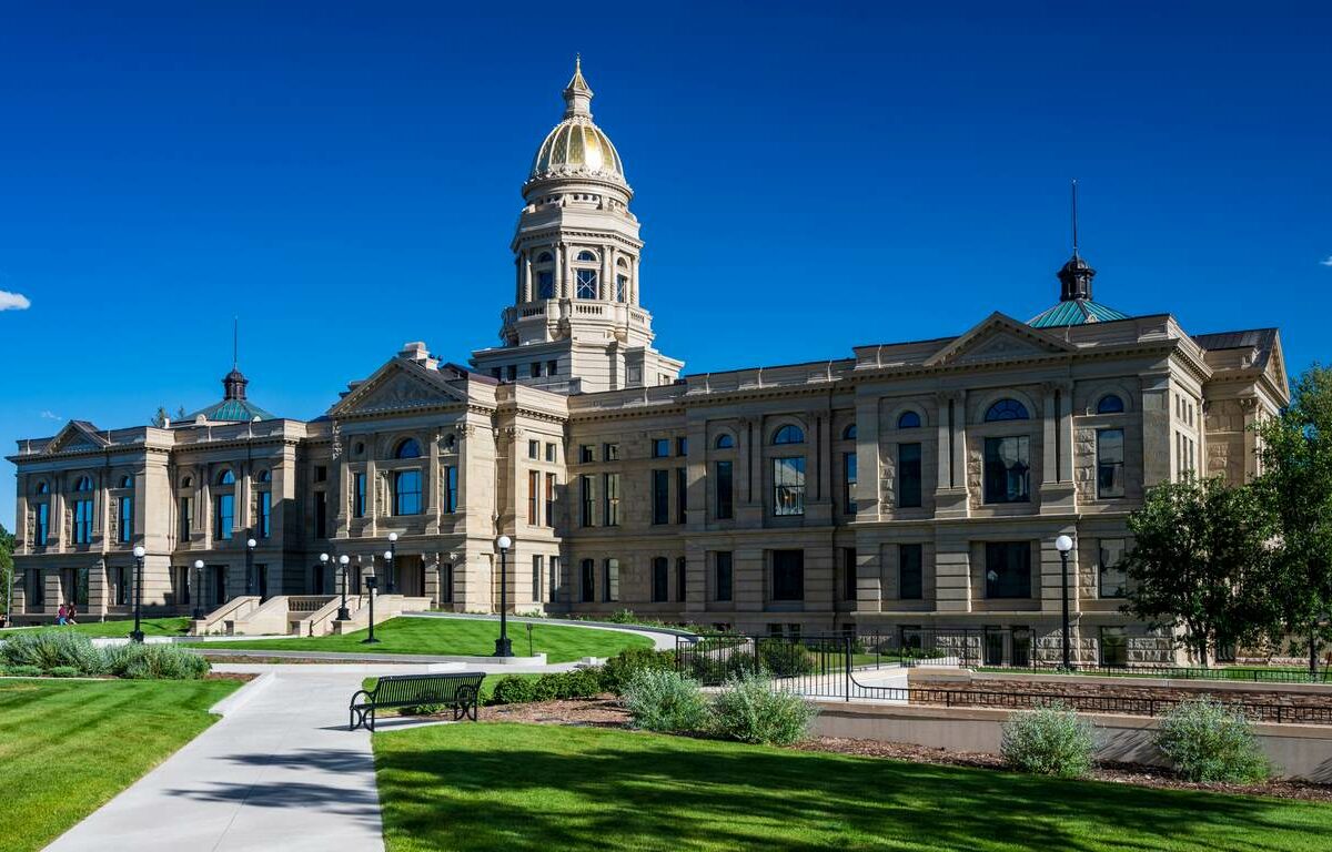 The Wyoming State Capitol, a large historic building with a gold dome, surrounded by a manicured lawn and blue skies.