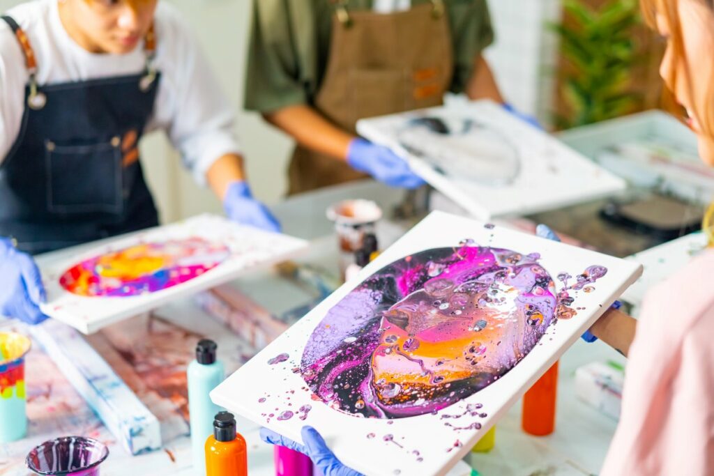 Multiple young people in an art workshop holding canvases covered with black, purple, orange, and pink acrylic paints.