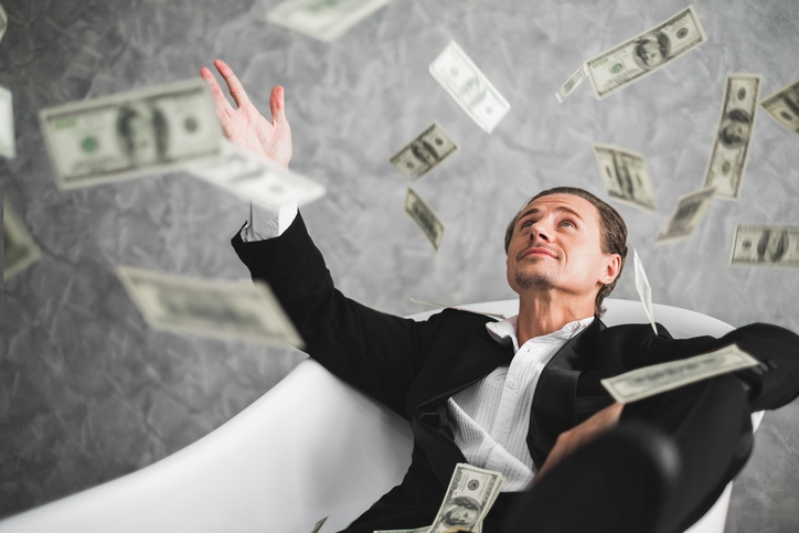 A man in a suit sits in an empty bathtub and smiles as cash falls from above.